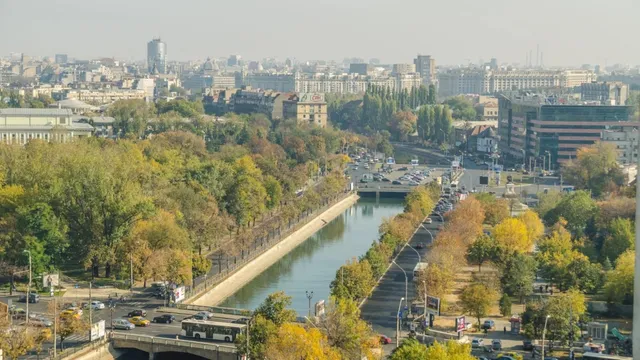 O vedere panoramică asupra unui oraș cu un canal central, copaci cu frunziș verde și galben, clădiri și mașini pe drumuri.