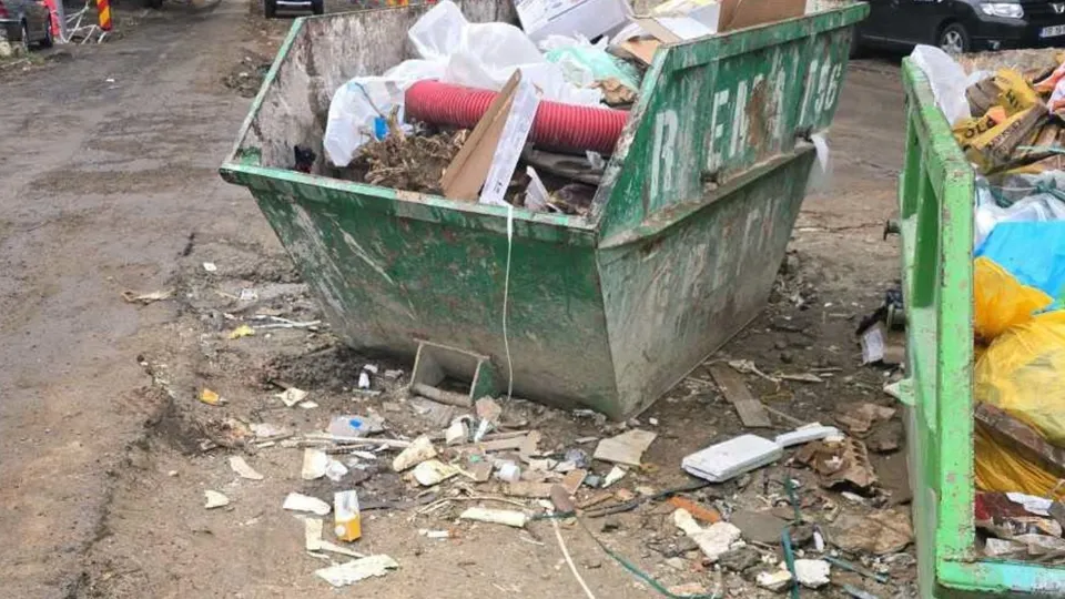 Un container verde metalic plin cu deșeuri și resturi de materiale de construcție este situat pe un teren de pământ murdar.