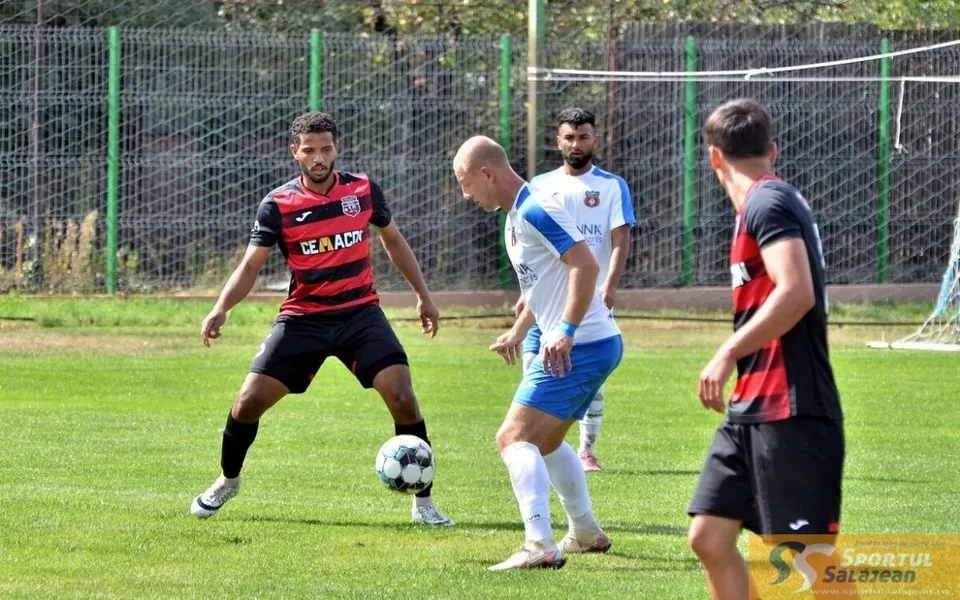 Trei jucători de fotbal sunt pe un teren de gazon verde, unul având un balon la picioare, în fața unui gard de sârmă.