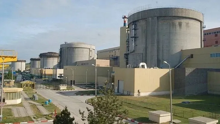 Două structuri cilindrice masive de beton gri sunt situate lângă clădiri industriale de culoare galbenă sub un cer senin.