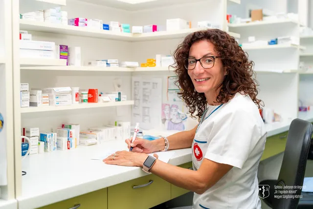 O femeie cu părul creț și ochelari scrie pe o foaie de hârtie de pe un blat alb, în fața unor rafturi cu cutii de medicamente.