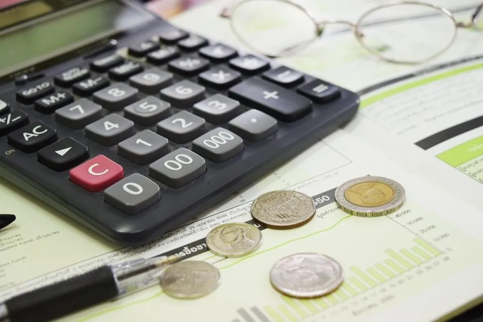 Un calculator negru, ochelari cu ochelari rotunzi, monede și un stilou sunt așezate pe documente cu grafice pe o suprafață deschisă.