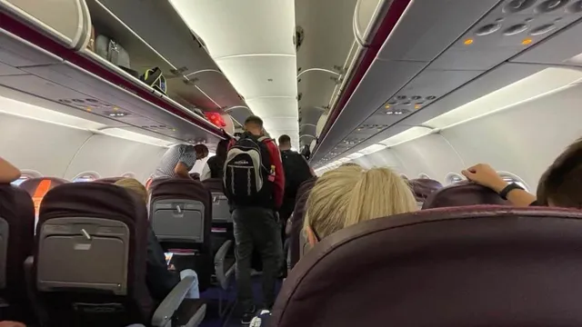 Pasageri stau așezați în scaunele unui avion, în timp ce un bărbat cu un rucsac negru merge pe culoarul dintre rândurile de scaune.