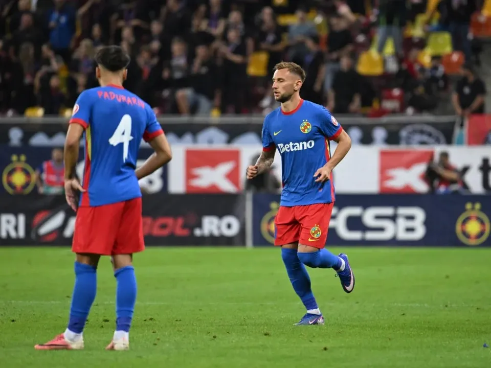 Doi jucători de fotbal în echipament albastru și roșu se află pe un teren de fotbal verde în fața unor tribune cu spectatori.