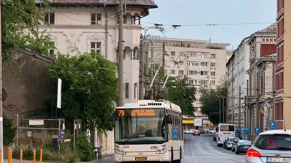 Un tramvai alb cu numărul 79 circulă pe o stradă urbană lângă clădiri înalte și mașini parcate.