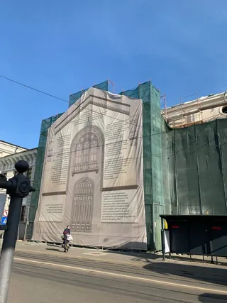 O persoană merge pe trotuar lângă un șantier acoperit de panouri verzi și un panou publicitar mare cu o schiță a unei clădiri.