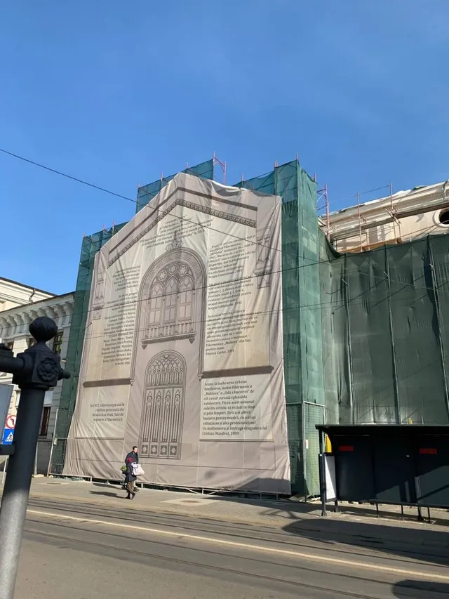 O persoană merge pe trotuar lângă un șantier acoperit de panouri verzi și un panou publicitar mare cu o schiță a unei clădiri.