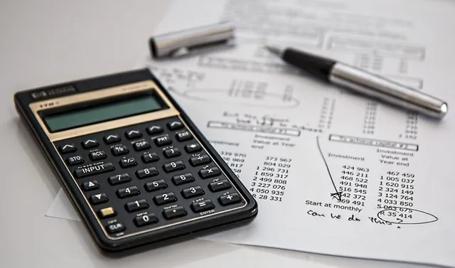 Un calculator negru, un stilou și un agrafator sunt așezate pe documente cu cifre și tabele pe o suprafață albă.