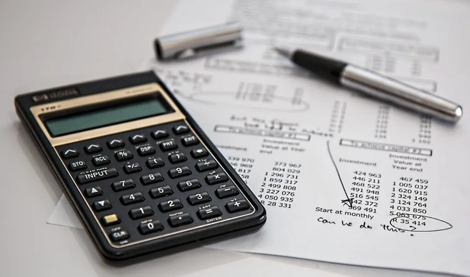 Un calculator negru, un stilou și un agrafator sunt așezate pe documente cu cifre și tabele pe o suprafață albă.
