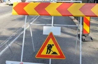 Un semn de circulație triunghiular cu margini roșii și pictograma unui om lucrând cu o lopată este montat pe un barieră metalică de șantier.