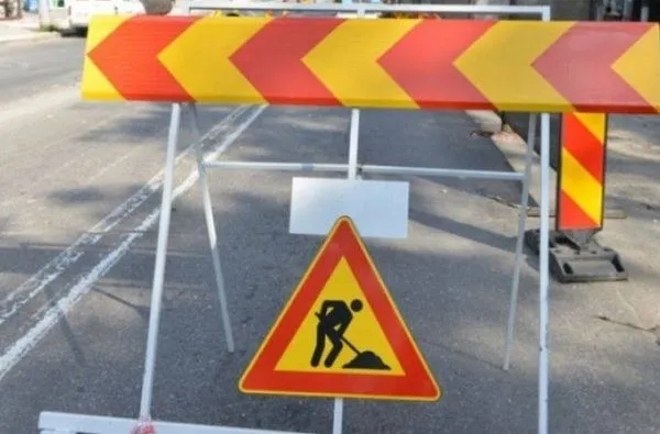 Un semn de circulație triunghiular cu margini roșii și pictograma unui om lucrând cu o lopată este montat pe un barieră metalică de șantier.