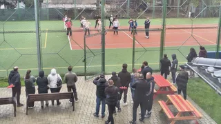 O grupă de persoane observă copii care practică activități sportive pe un teren de tenis / sport în aer liber, dintr-un unghi îndepărtat, prin gard.