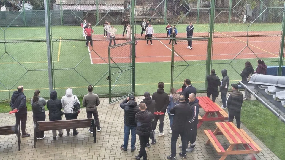O grupă de persoane observă copii care practică activități sportive pe un teren de tenis / sport în aer liber, dintr-un unghi îndepărtat, prin gard.