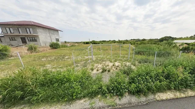 Un teren cu vegetație verde este delimitat de un gard de sârmă, având o clădire gri cu acoperiș roșu în fundal.