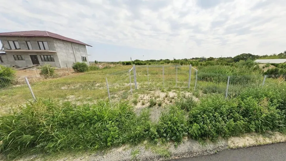 Un teren cu vegetație verde este delimitat de un gard de sârmă, având o clădire gri cu acoperiș roșu în fundal.