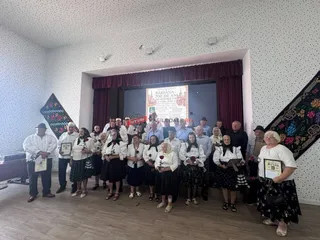 Un grup de persoane în haine tradiționale stau în rând pe o scenă în fața unui panou decorativ cu text.