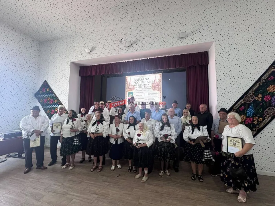 Un grup de persoane în haine tradiționale stau în rând pe o scenă în fața unui panou decorativ cu text.
