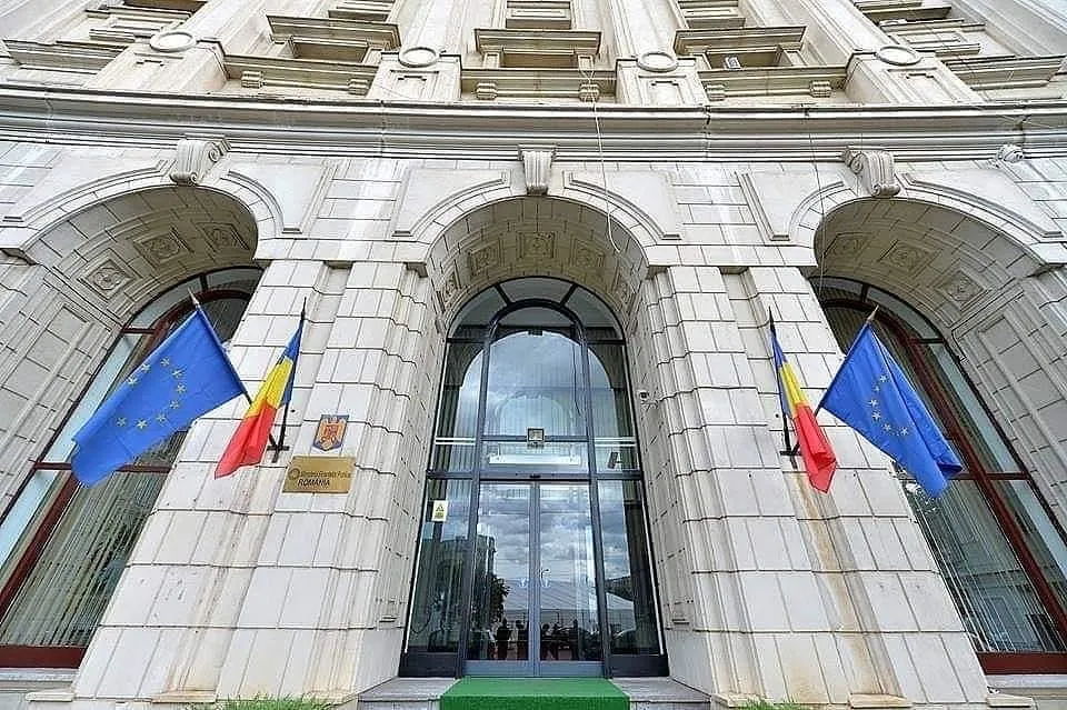 O clădire de piatră cu trei arcuri mari prezintă trei steaguri și o intrare cu uși de sticlă.