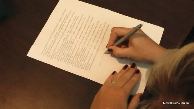O persoană scrie cu un pix pe o foaie de hârtie albă aflată pe o masă închisă la culoare.