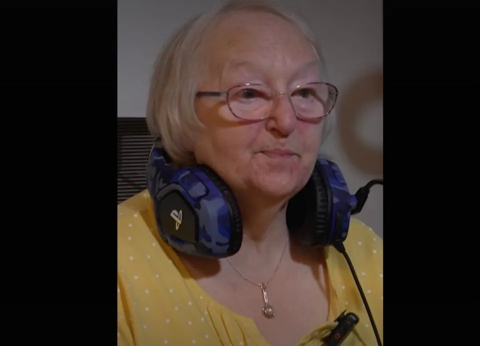 O femeie cu ochelari și căști de gaming albastre poartă un colier cu pandantiv și un tricou galben cu puncte albe.