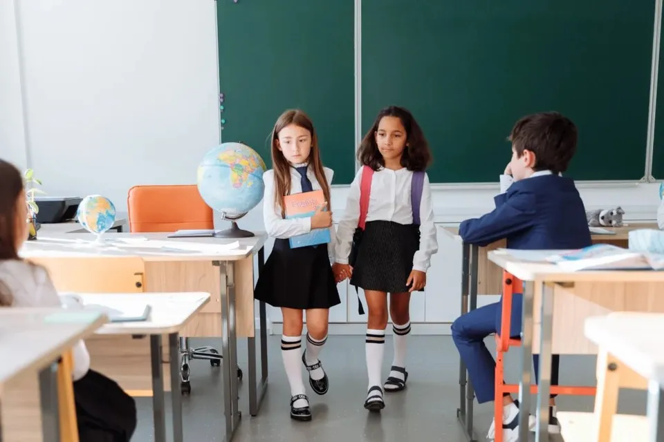 Două fete în uniformă școlară merg printre bănci, în timp ce un băiat stă așezat la o masă într-o sală de clasă.