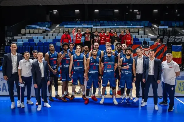 Un grup de baschetbaliști și bărbați în costume pozează pe parchet, unii ridicând trofee, în fața tribunelor unei săli de sport.