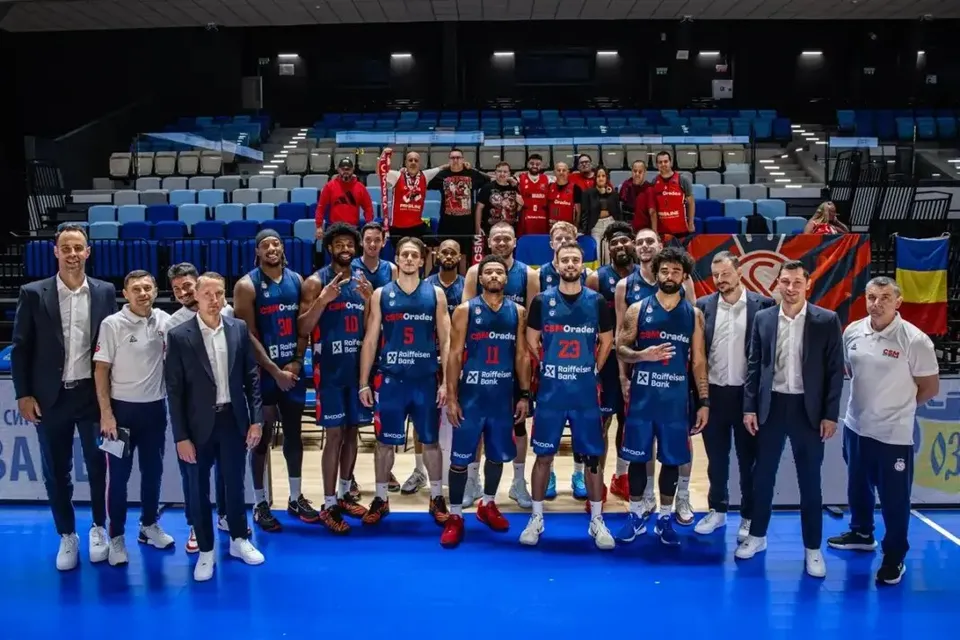 Un grup de baschetbaliști și bărbați în costume pozează pe parchet, unii ridicând trofee, în fața tribunelor unei săli de sport.