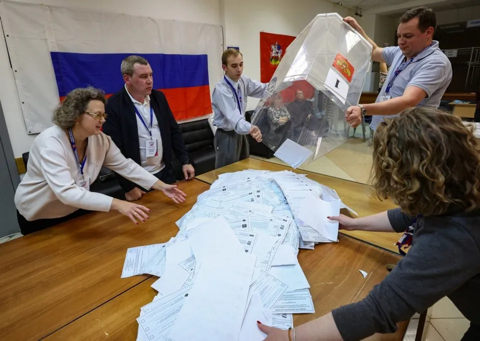 Cinci persoane verifică și manipulează buletine de vot pe o masă, în prezența drapelului rus.