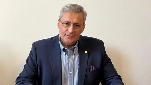 Un bărbat cu ochelari, purtând un sacou albastru și o cămașă cu dungi, stă în fața unui fundal alb.