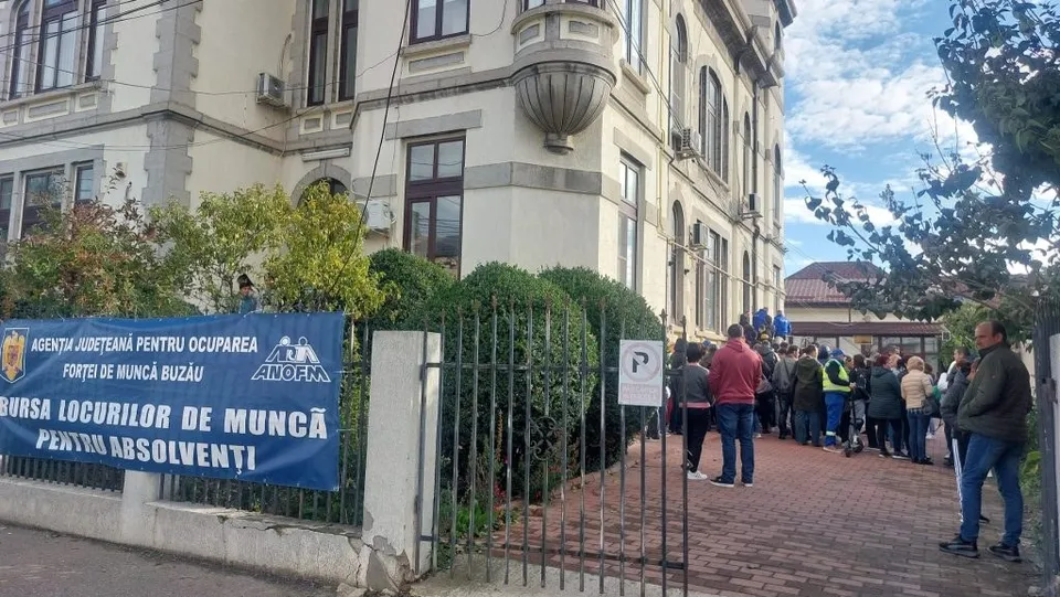 Oamenii stau la coadă pe un trotuar lângă o clădire cu un banner albastru pe care scrie text alb.