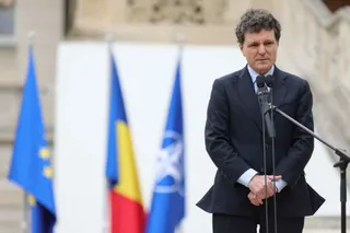 Un bărbat cu părul încrețit, îmbrăcat într-un costum închis la culoare, stă în fața unor microfoane și a unor steaguri colorate.