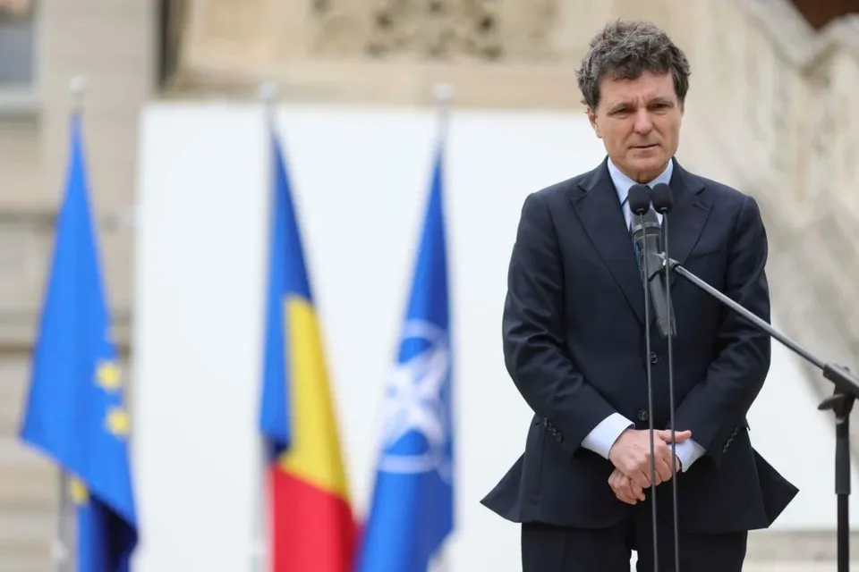 Un bărbat cu părul încrețit, îmbrăcat într-un costum închis la culoare, stă în fața unor microfoane și a unor steaguri colorate.