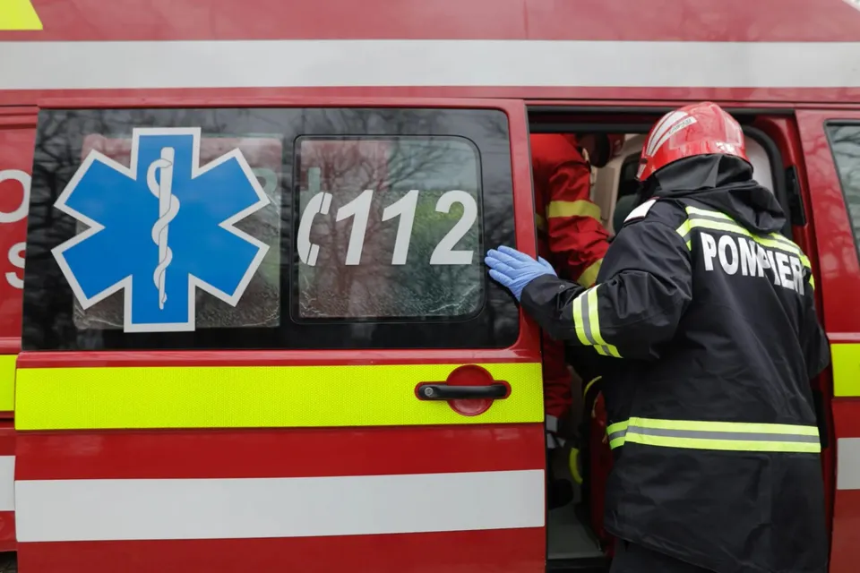 Un pompier cu cască roșie și uniformă neagră stă lângă o ambulanță roșie cu simbolul Star of Life și numărul 112 pe fereastră.
