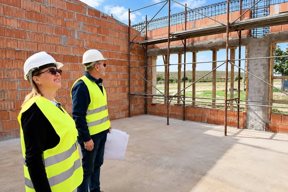 Două persoane purtând căști albe și veste reflectorizante galben-verzi stau într-o clădire în construcție cu pereți din cărămidă și schele metalice.