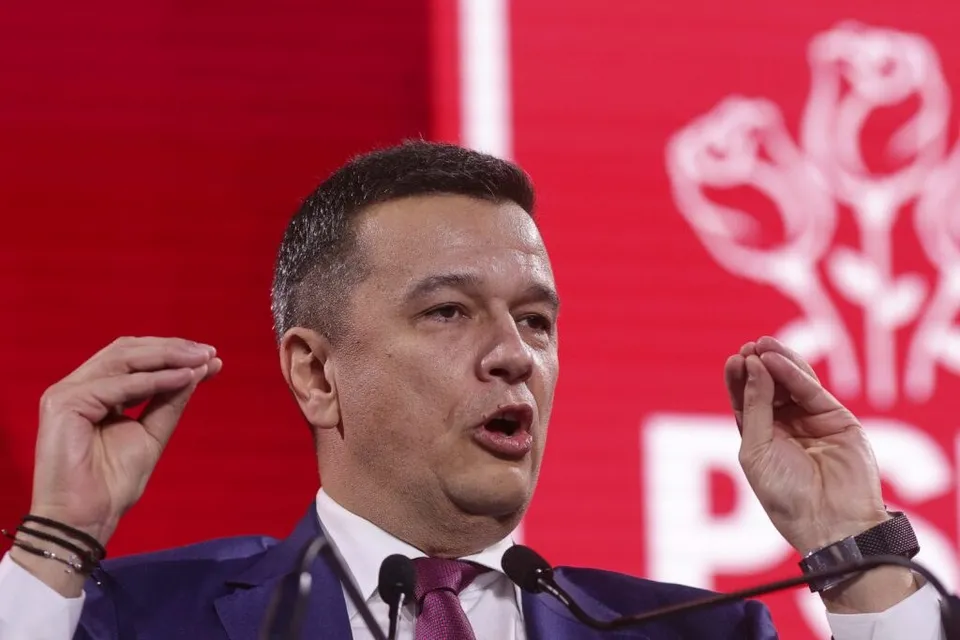 Un bărbat cu părul scurt, purtând sacou albastru și cămașă albă, ridică ambele mâini cu degetele apropiate în poziție de gestuală în fața unor microfoane.