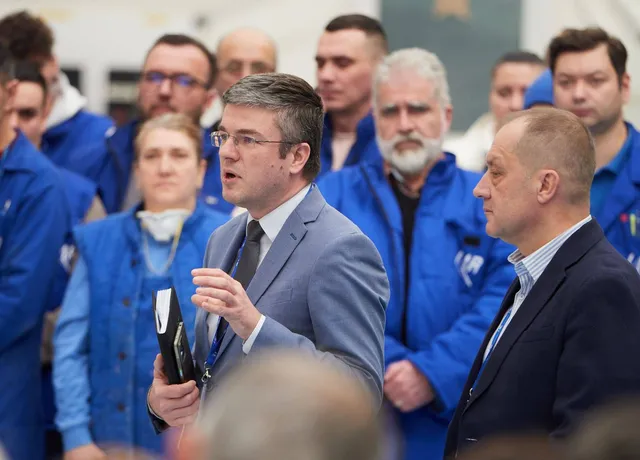 Un bărbat în costum gri cu cravată vorbește, înconjurat de persoane în uniforme albastre și alți bărbați în costume.