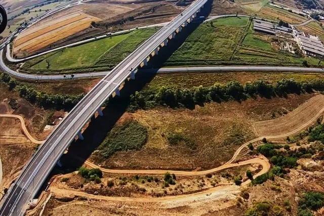 O vedere aeriană prezintă un viaduct lung de asfalt care traversează un peisaj rural format din câmpuri agricole, vegetație și drumuri de pământ.