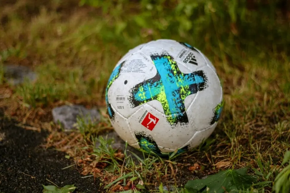 Un fotbal o albă cu modele albastre și negre este așezată pe iarbă lângă o porțiune de asfalt.