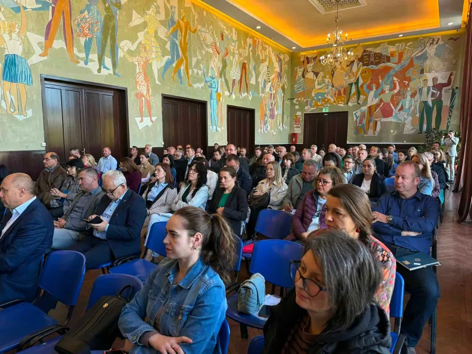 Un grup de persoane stă așezat pe scaune albastre într-o sală cu pereți decorați cu un mural colorat cu figuri umane.