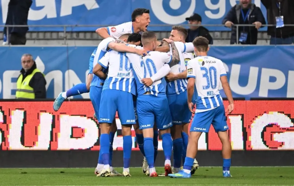 Un grup de fotbaliști în echipament albastru și alb se îmbrățișează pe un teren de fotbal în fața unui panou publicitar luminos.