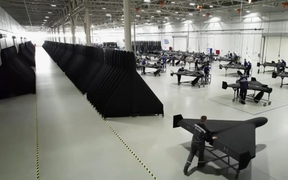 Oamenii lucrează într-o fabrică mare, unde sunt așezate pe podea numeroase componente negre de formă aerodinamică și mese de lucru cu unelte.