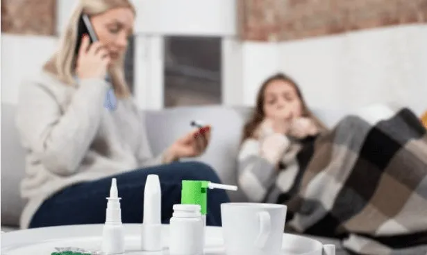 O femeie vorbește la telefon, în timp ce o altă persoană stă în spatele ei sub o pătură, având diverse flacoane și o cană pe o masă.
