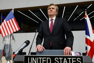 Un bărbat într-un costum închis la culoare și cravată roșie stă în spatele unui podium cu inscripția UNITED STATES, între două steaguri.