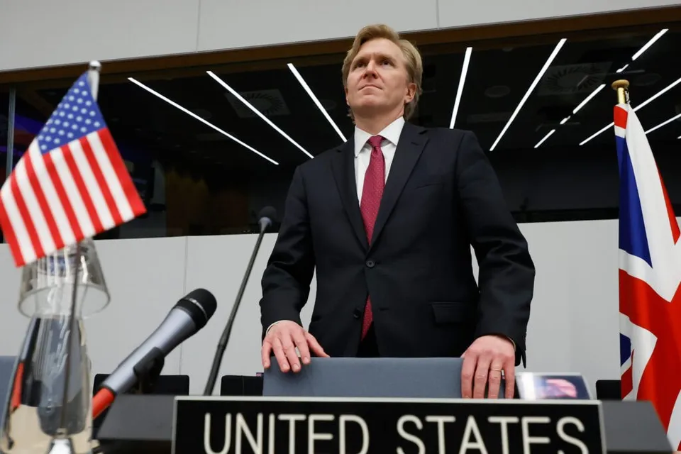 Un bărbat într-un costum închis la culoare și cravată roșie stă în spatele unui podium cu inscripția UNITED STATES, între două steaguri.