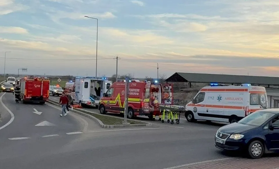 Mai multe vehicule de serviciu, inclusiv ambulanțe și o autospecială de pompieri, sunt parcate pe o porțiune de drum asfaltat sub un cer înnorat.