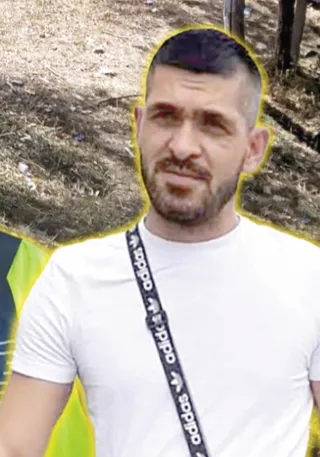 Un bărbat cu barbă și păr scurt poartă un tricou alb și un cordon negru cu logo-uri albe peste umăr.