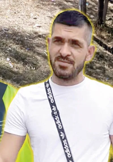 Un bărbat cu barbă și păr scurt poartă un tricou alb și un cordon negru cu logo-uri albe peste umăr.