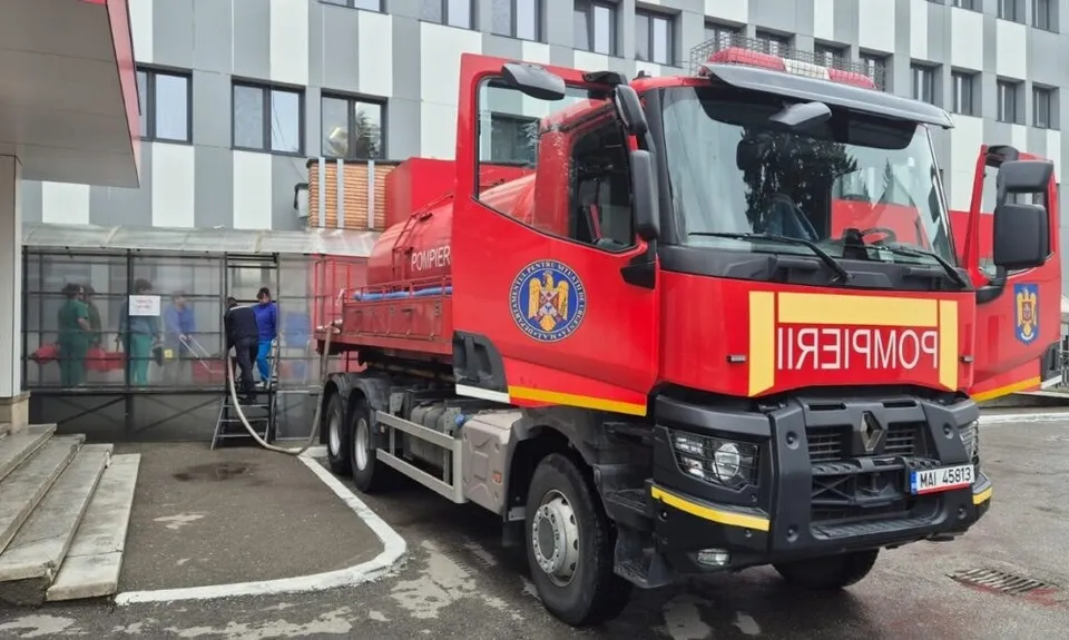 Un camion de pompieri roșu este parcat în fața unei clădiri gri cu ferestre de sticlă.