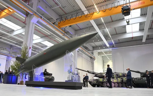 Un obiect aerodinam de culoare verde înclinat este expus într-un hangar mare lângă vehicule militare și persoane care privesc.