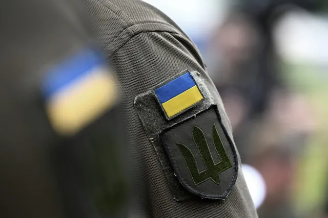 Un patch dreptunghiular albastru și galben este cusut pe mâneca unei uniforme de culoare verde măsliniu, sub un alt patch negru cu un simbol trident.
